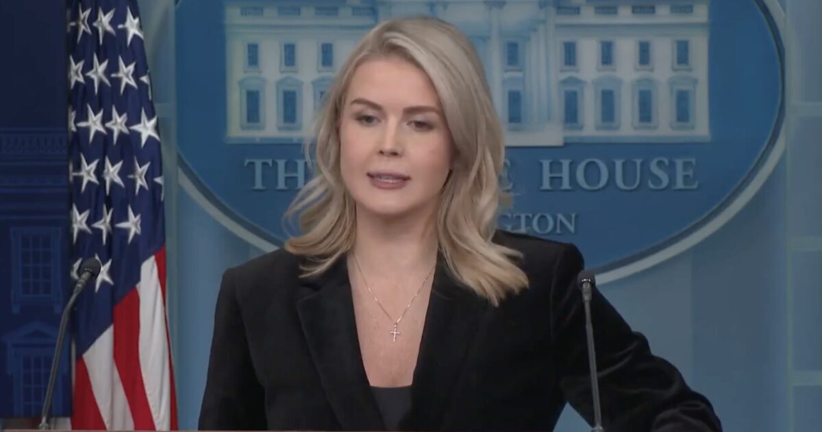 Press secretary delivering a statement at the White House briefing room, with the American flag in the background.